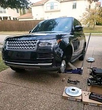 Un Range Rover negro est&aacute; estacionado en la entrada de una vivienda, con la rueda delantera del lado del conductor desmontada. Hay un gato debajo del veh&iacute;culo y varias piezas de repuesto esparcidas por la entrada.